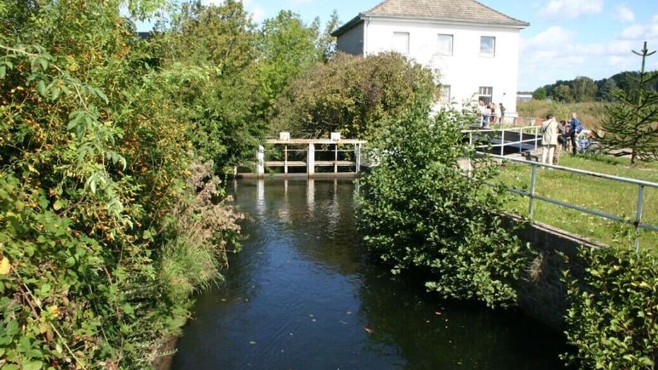 A narrow waterway flanked by dense foliage, a wooden bridge, and a white building in the background.