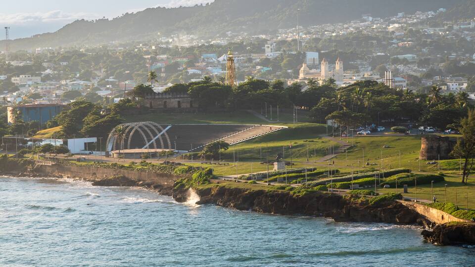 A coastal cityscape with a prominent clock tower, a stadium, and a rocky shoreline.