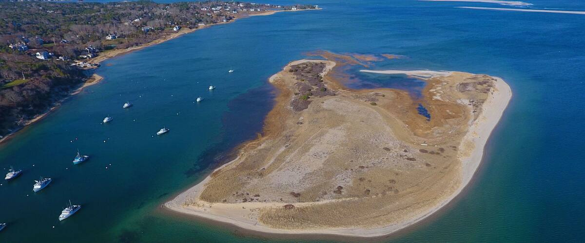 An aerial view of a coastal area with a small island surrounded by clear blue waters. There are several boats anchored near the shore, and a dense forested area is visible to the left.