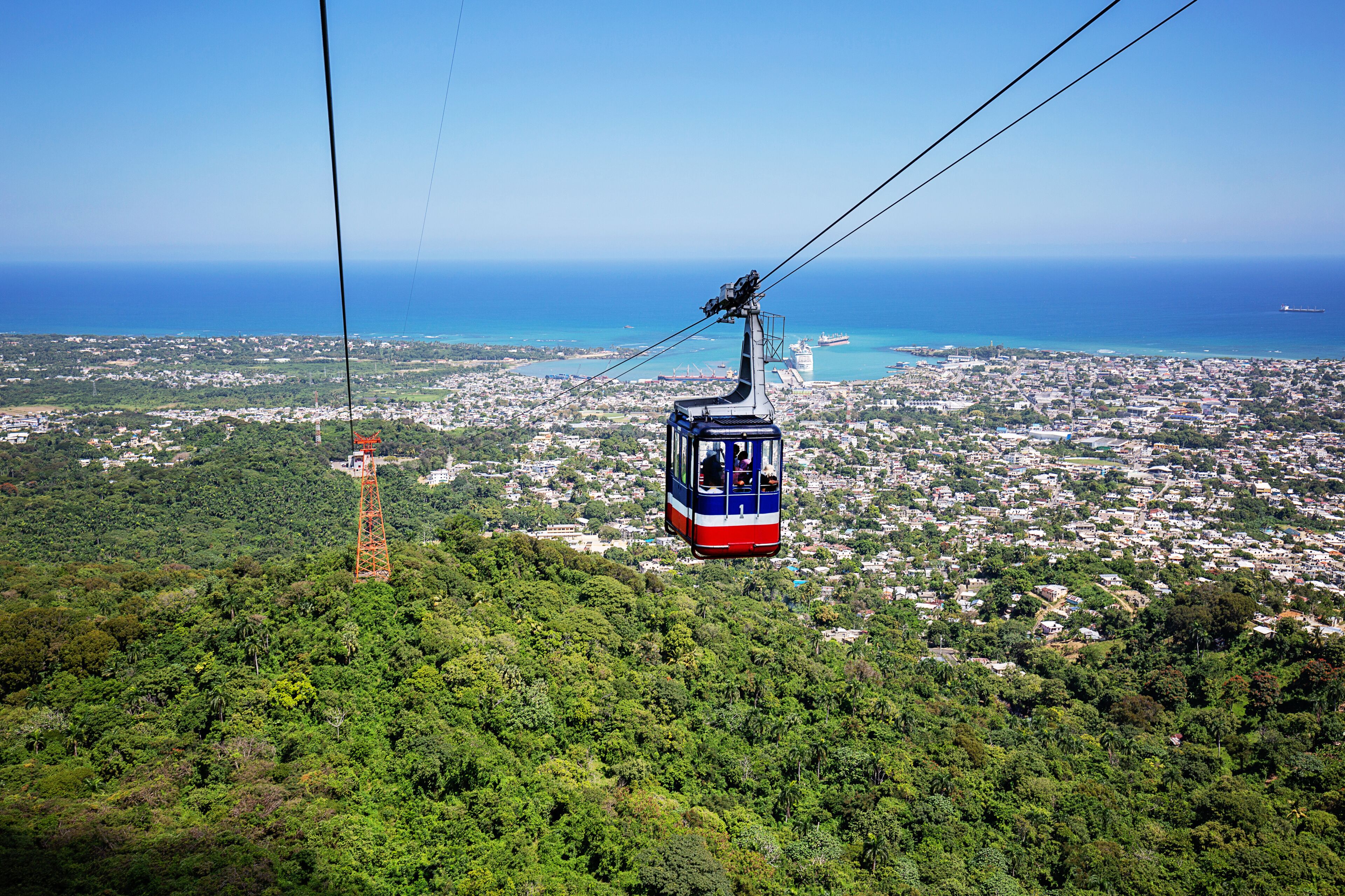 A cable car over a densely populated city with a view of the ocean and a ship in the distance.