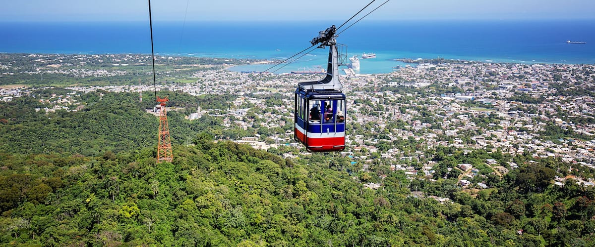 A cable car over a densely populated city with a view of the ocean and a ship in the distance.