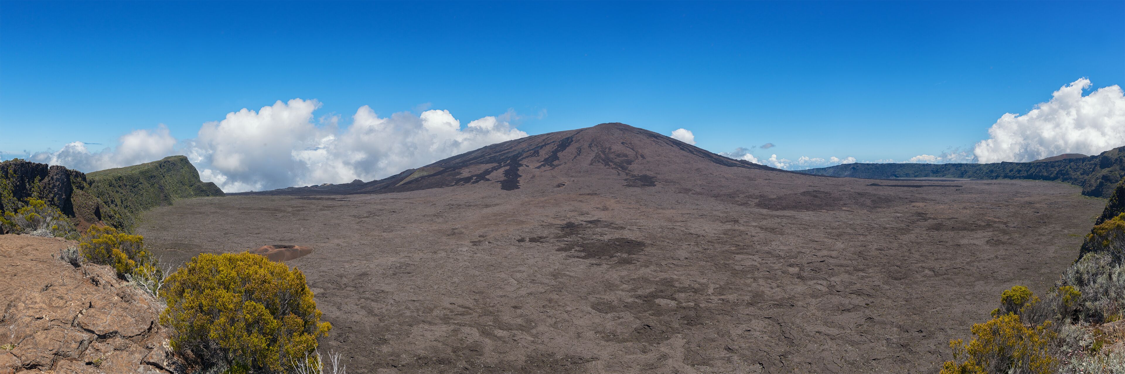 A barren volcanic landscape with a prominent mountain in the center, surrounded by sparse vegetation and a clear blue sky.