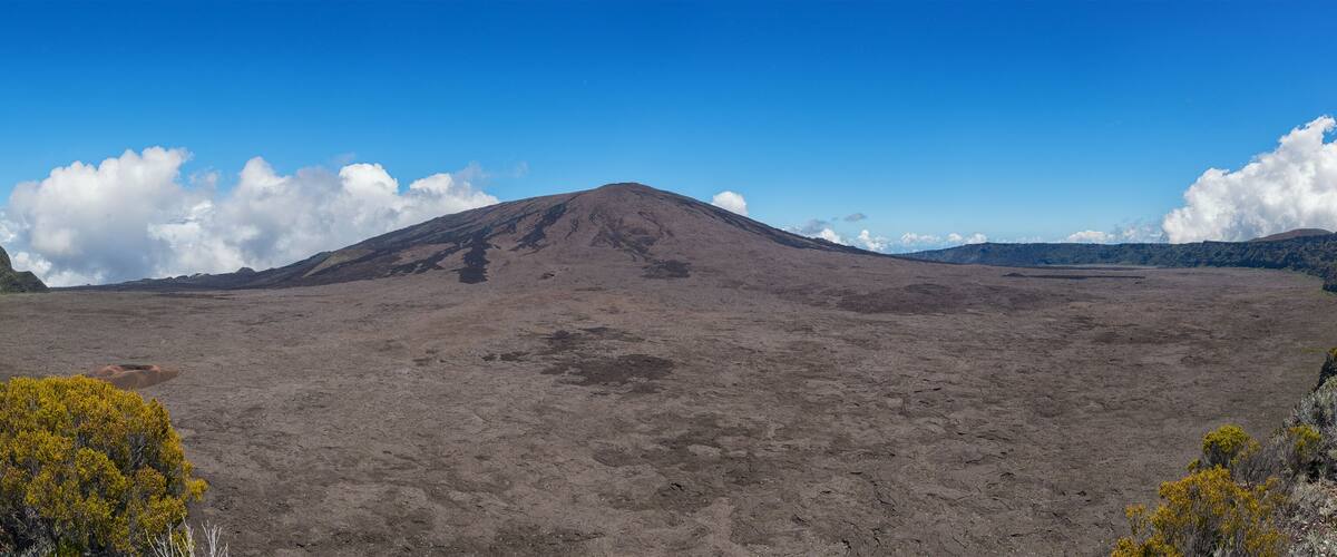 A barren volcanic landscape with a prominent mountain in the center, surrounded by sparse vegetation and a clear blue sky.