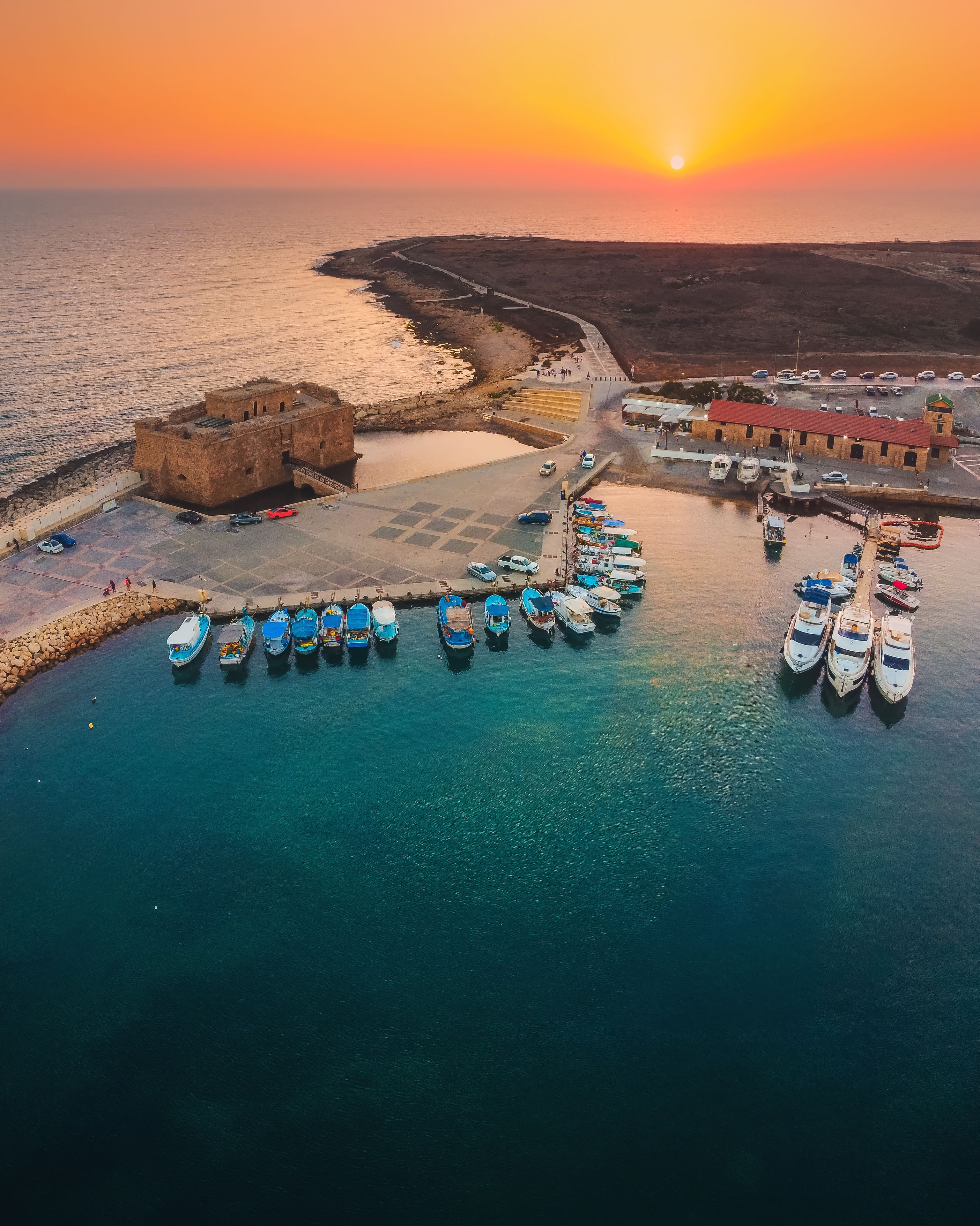 A coastal harbor at sunset with boats docked, a fortified structure, and a pier extending into the water.