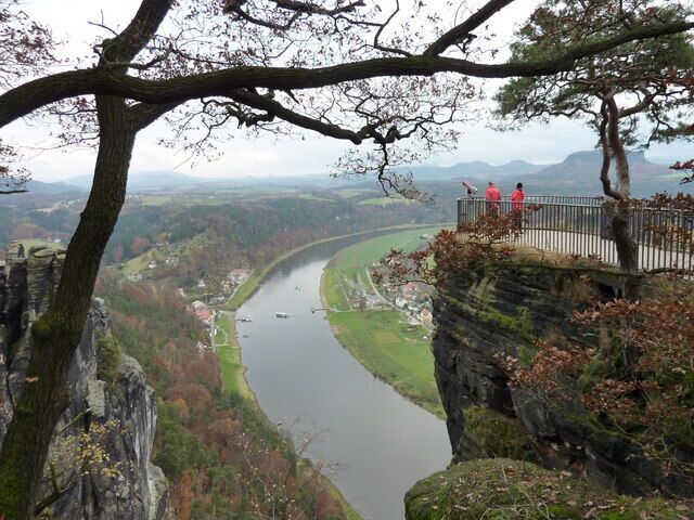 Ein Fluss schlängelt sich durch ein Tal, eine Klippe mit Geländer und ein fernes Gebirge.