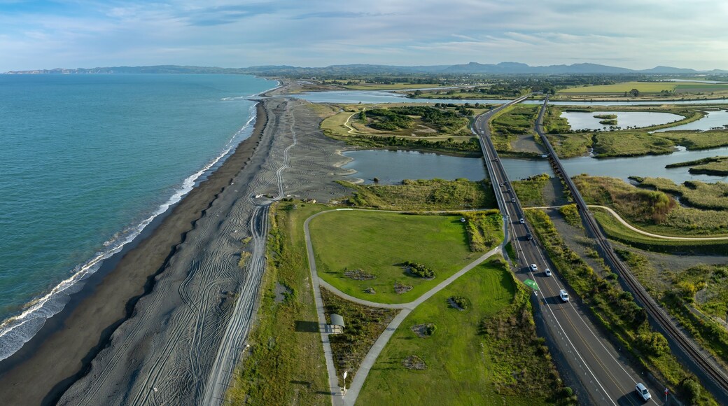Coastal landscape with a road, bridge, and estuary. Cars travel on the road, while the beach and surrounding wetlands are visible. Waitangi Regional Park, Napier, Hawke's Bay, New Zealand