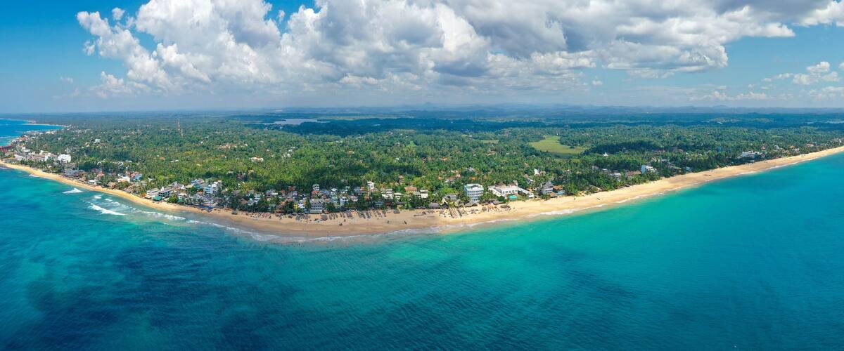 An aerial view of a coastal island with a beach, dense vegetation, and a clear blue sea.