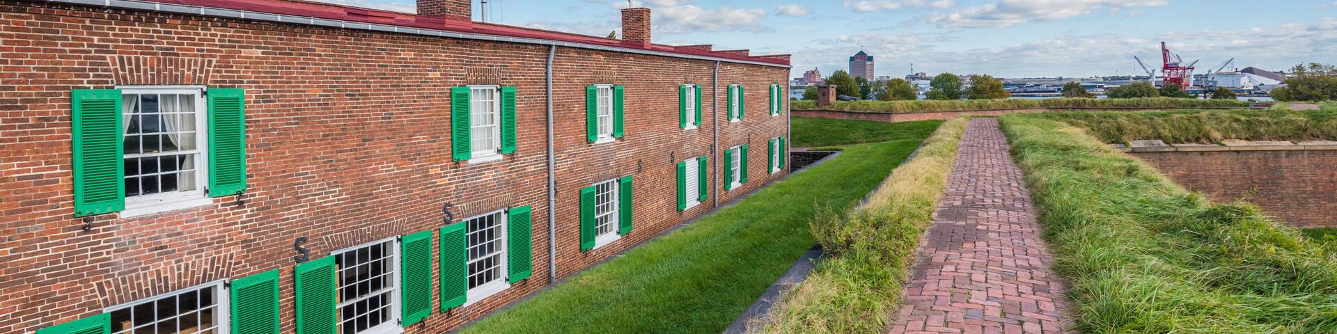 A brick building with green shutters and a flagpole, a brick pathway, and a clear blue sky with clouds.