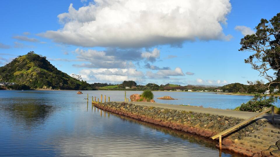 A calm lake with a wooden dock, a stone wall, and a hill with trees in the background.