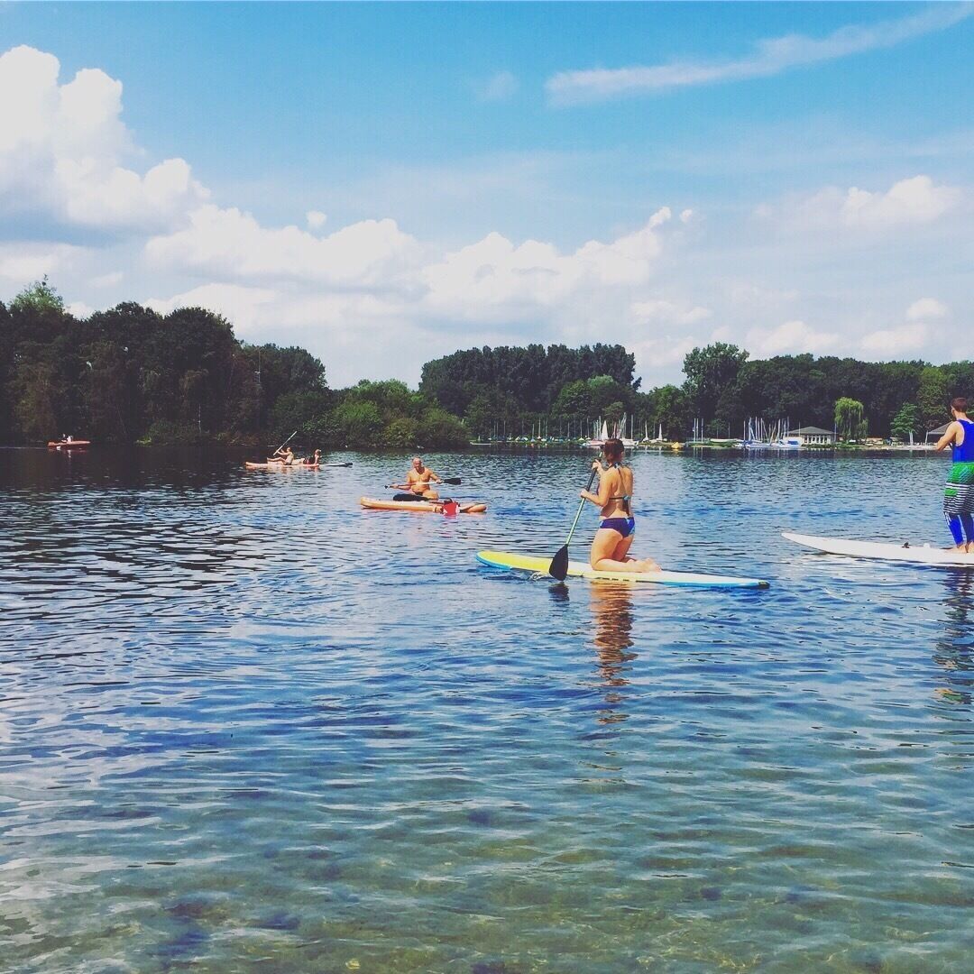People paddleboarding on a calm lake with clear water, surrounded by trees and boats in the background.