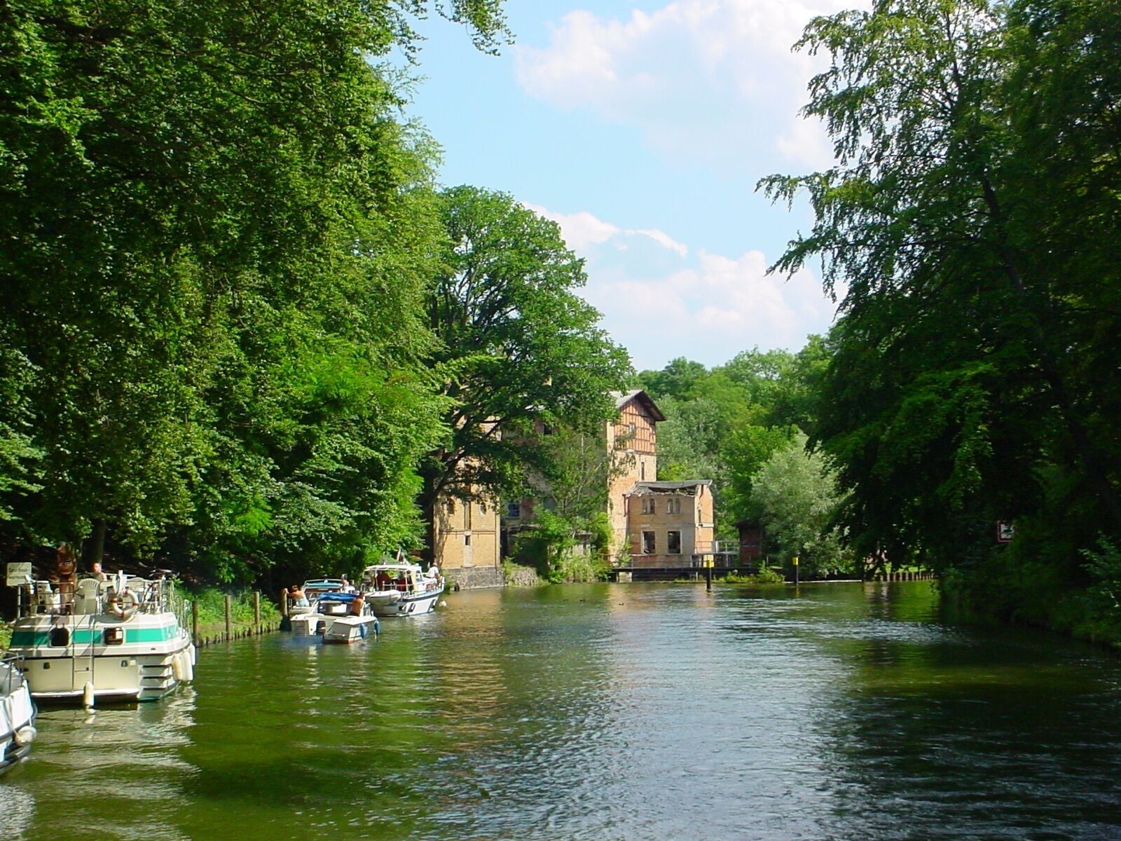 A river with boats, surrounded by lush green trees and a historic building with a tower.
