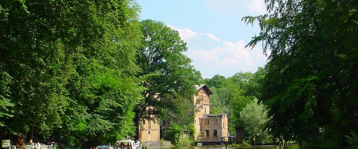 A river with boats, surrounded by lush green trees and a historic building with a tower.