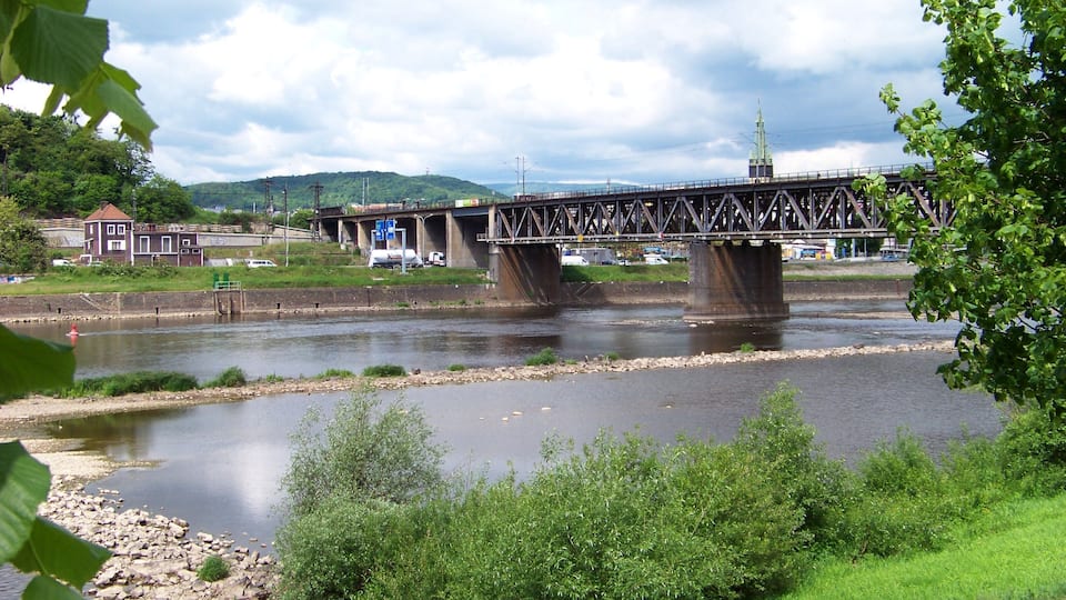 A steel bridge over a river with a building and vehicles on the opposite bank.