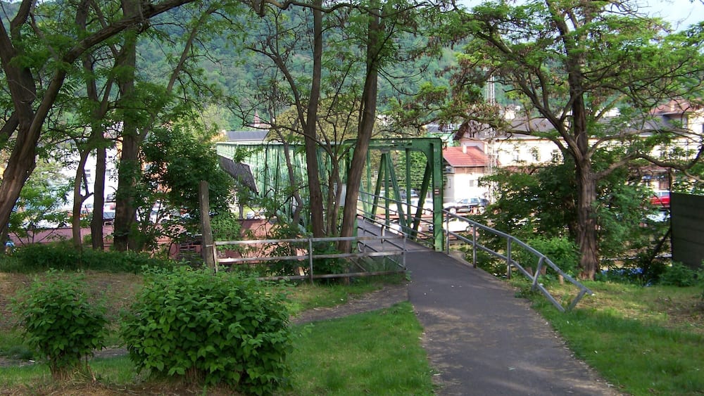 A green metal bridge over a paved path surrounded by trees and grass.