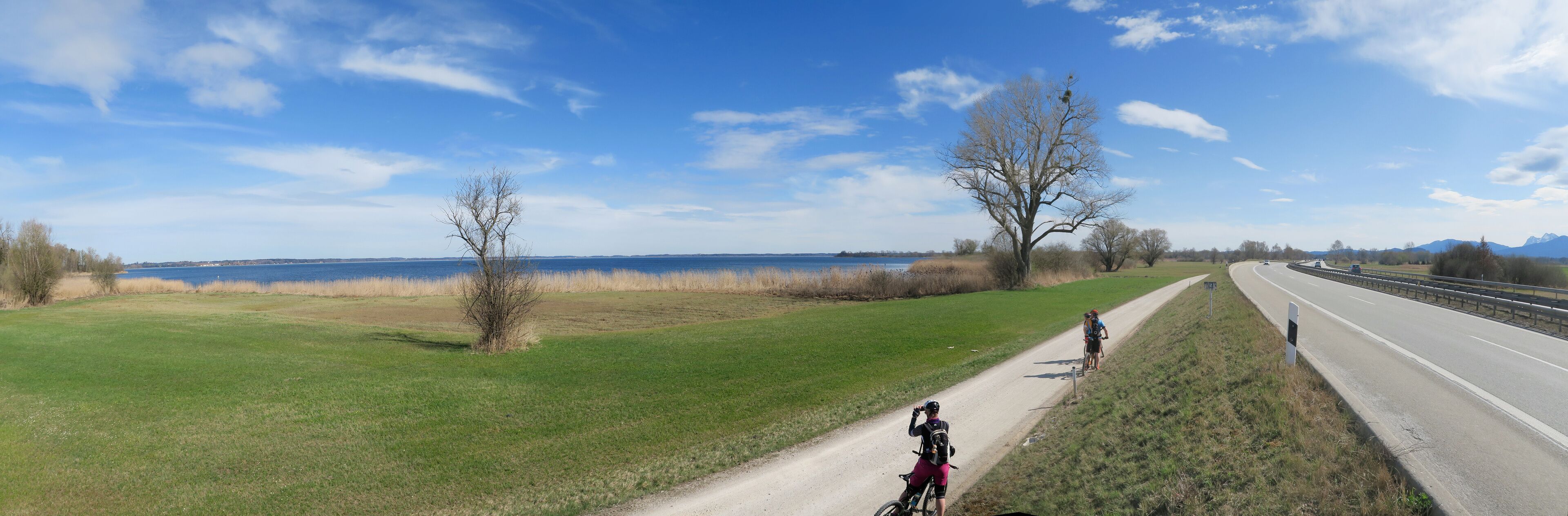 A paved path alongside a highway, with cyclists and a clear view of a lake and mountains in the distance.