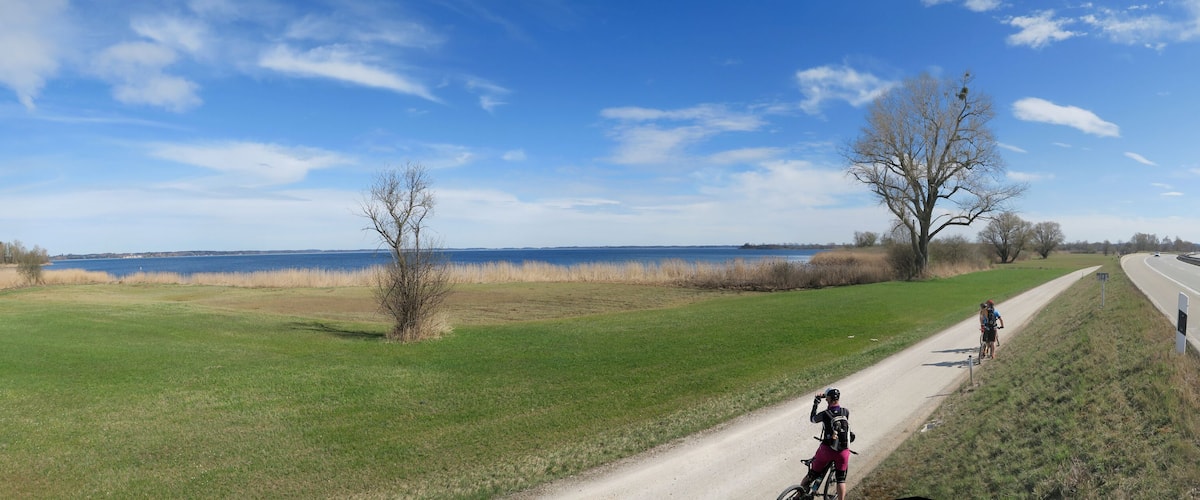 A paved path alongside a highway, with cyclists and a clear view of a lake and mountains in the distance.