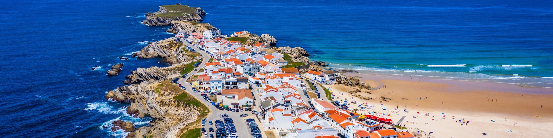 Un pueblo costero con playa, costa rocosa y edificios de tejados rojos.