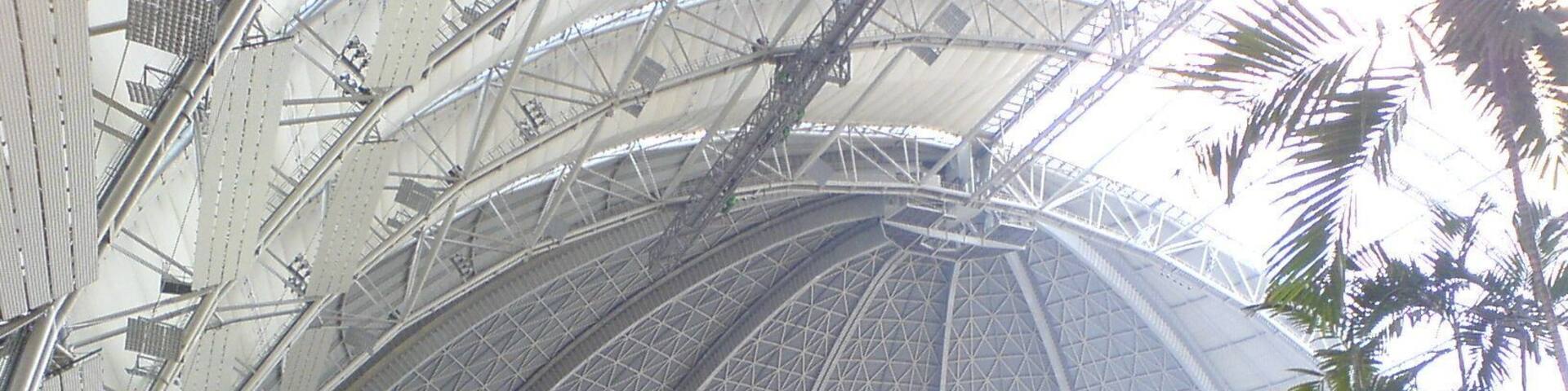 A large, domed structure with a metal framework and a thatched-roof pavilion in the foreground.