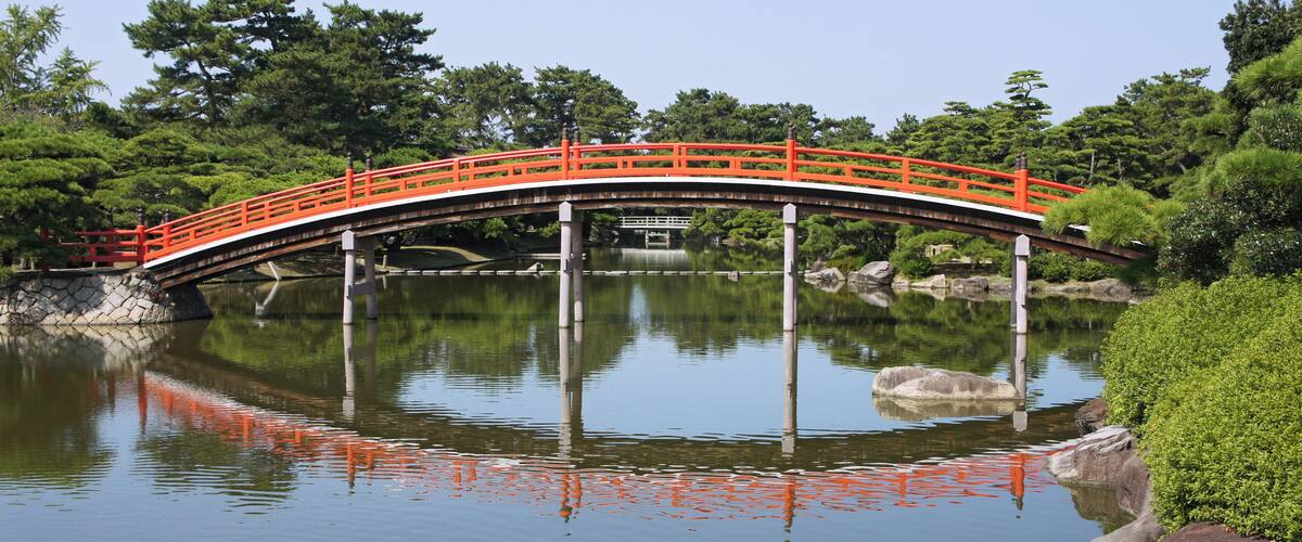 A red bridge over a calm pond with reflections, surrounded by lush greenery.