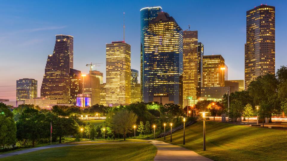 A city skyline with illuminated skyscrapers, a park with a pathway, and a body of water in the background.