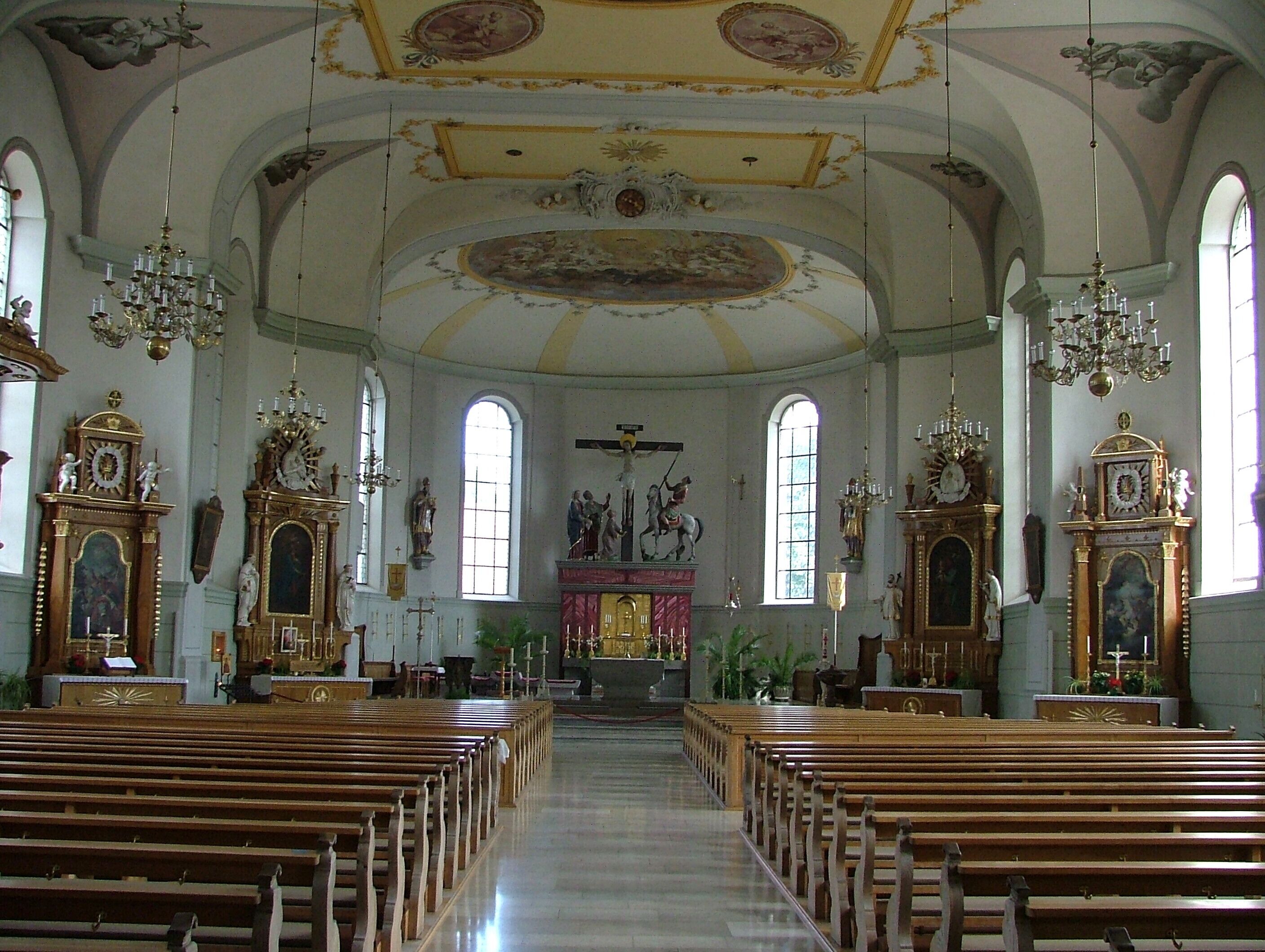 A church interior with wooden pews, chandeliers, and religious statues.