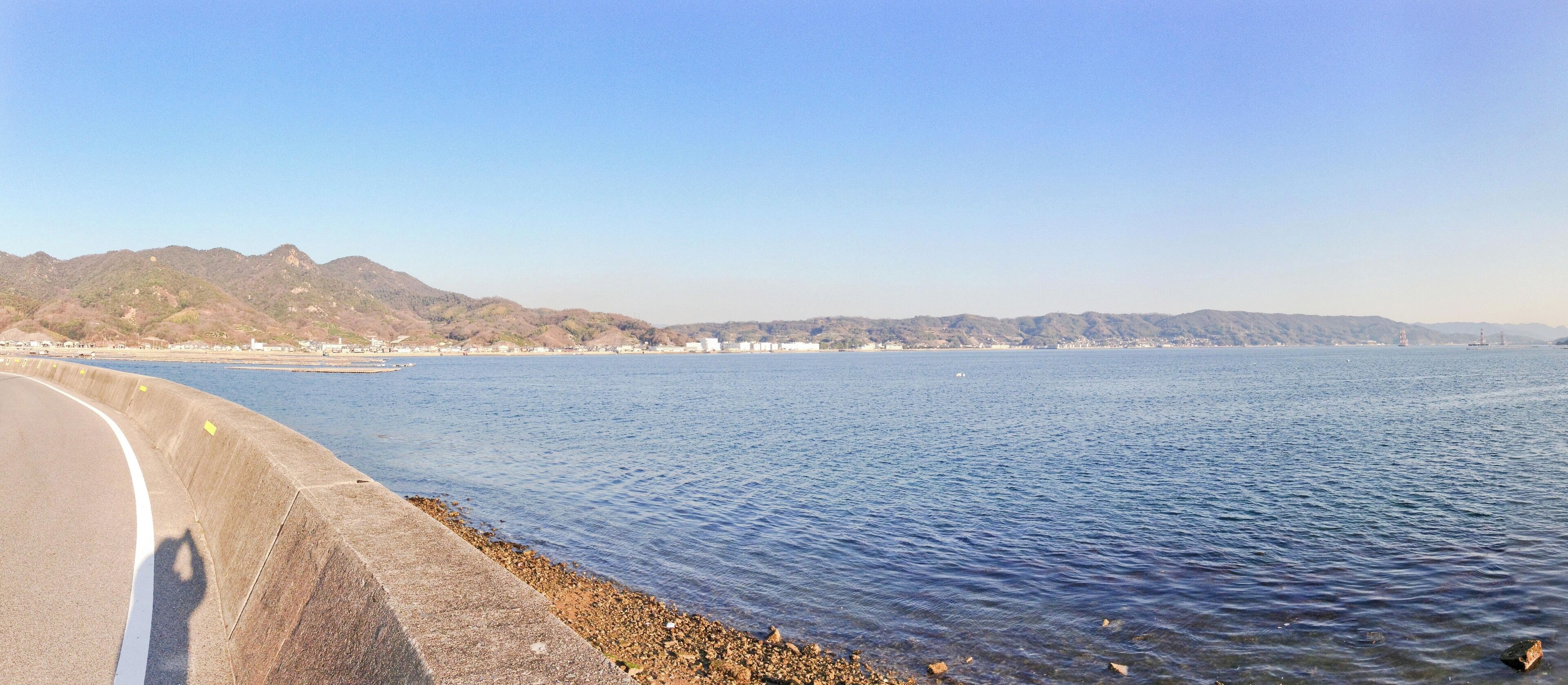 A coastal road with a concrete barrier, calm sea, and distant mountains.