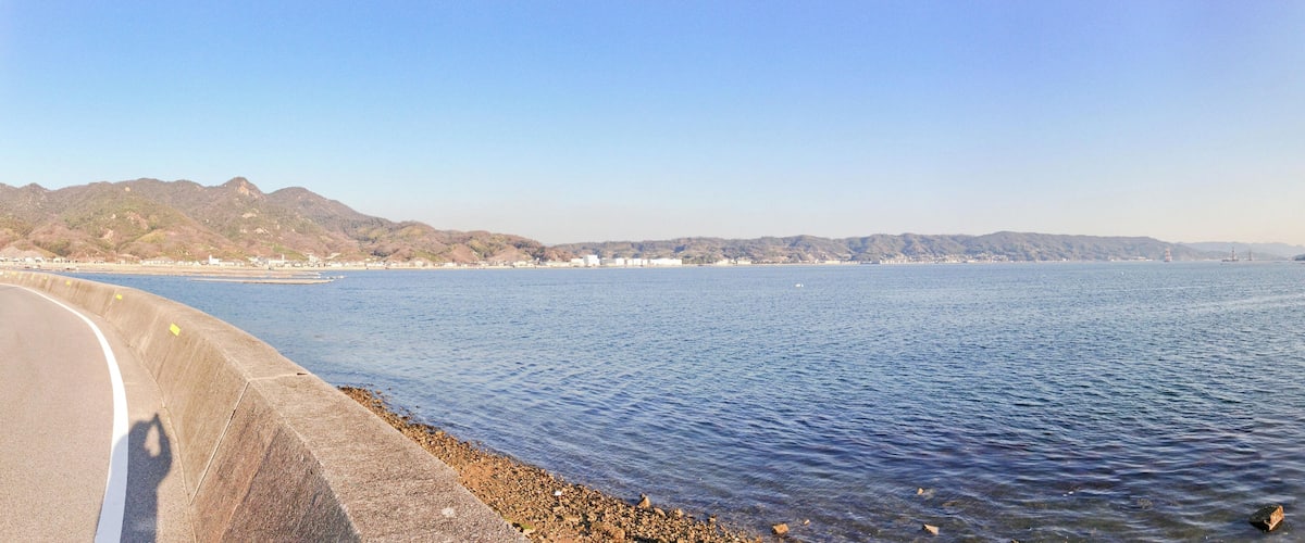 A coastal road with a concrete barrier, calm sea, and distant mountains.