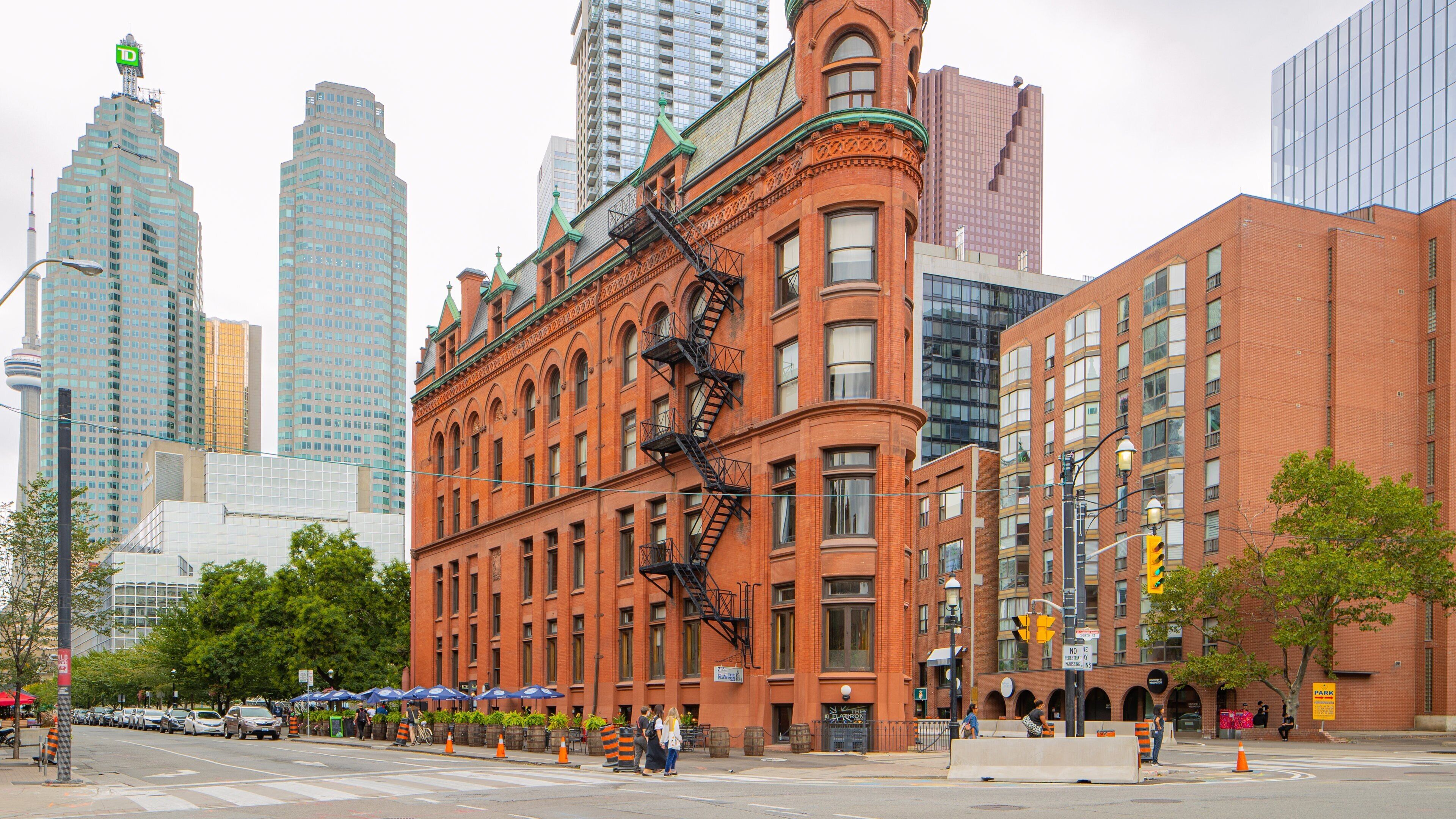 A historic red brick building with a green roof and ornate details, surrounded by modern skyscrapers and a busy street with pedestrians and parked cars.