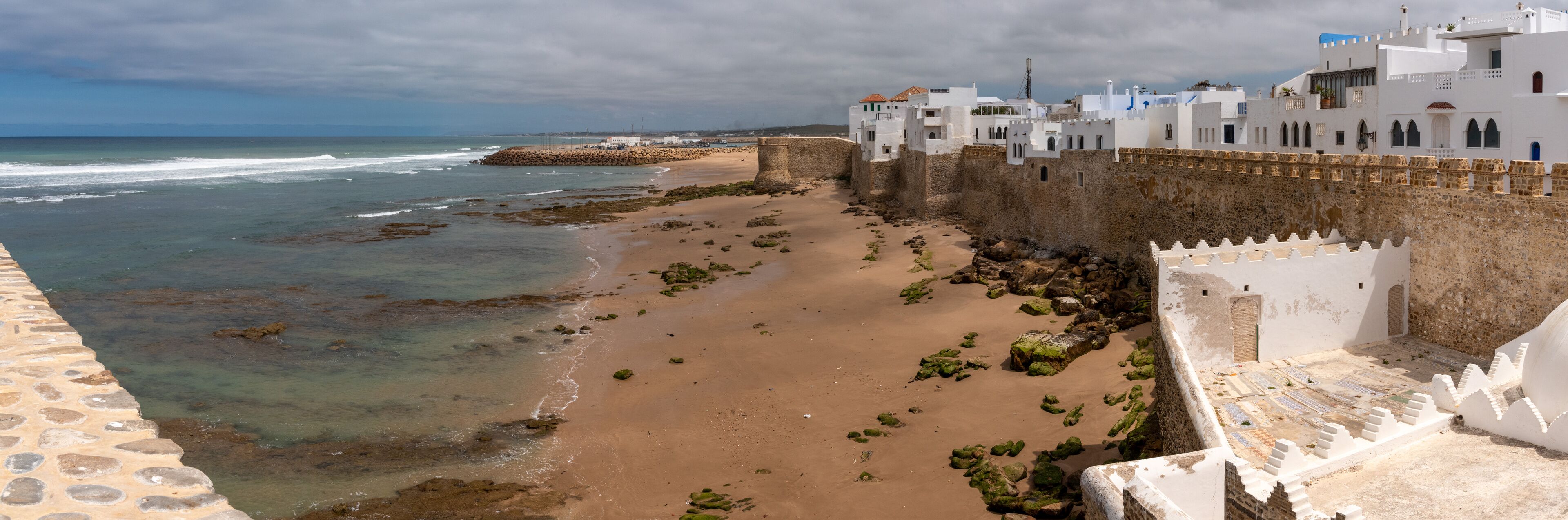 A coastal town with a sandy beach, a stone wall, and white buildings with blue accents.