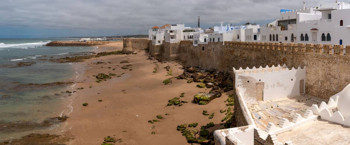 A coastal town with a sandy beach, a stone wall, and white buildings with blue accents.