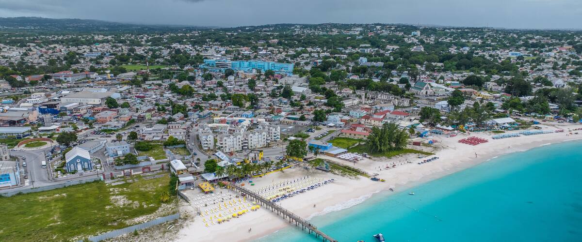An aerial view of a coastal city with a beach, a pier, and a dense urban area with colorful buildings.