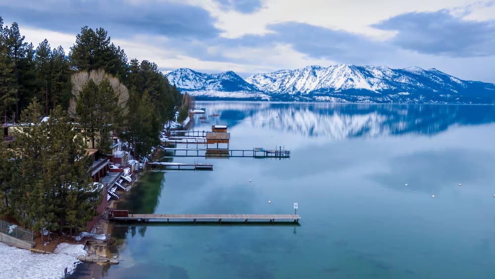 A calm lake with snow-capped mountains in the background, a wooden dock extending into the water, and a forested shoreline.