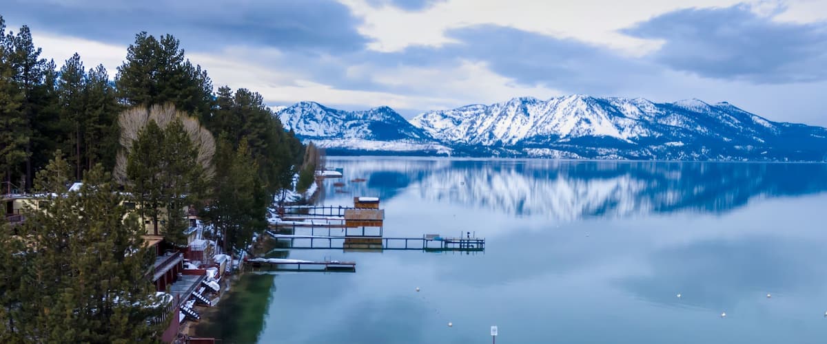 A calm lake with snow-capped mountains in the background, a wooden dock extending into the water, and a forested shoreline.