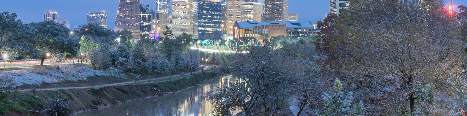 A city skyline with tall buildings, a river reflecting the lights, and trees with snow on branches.