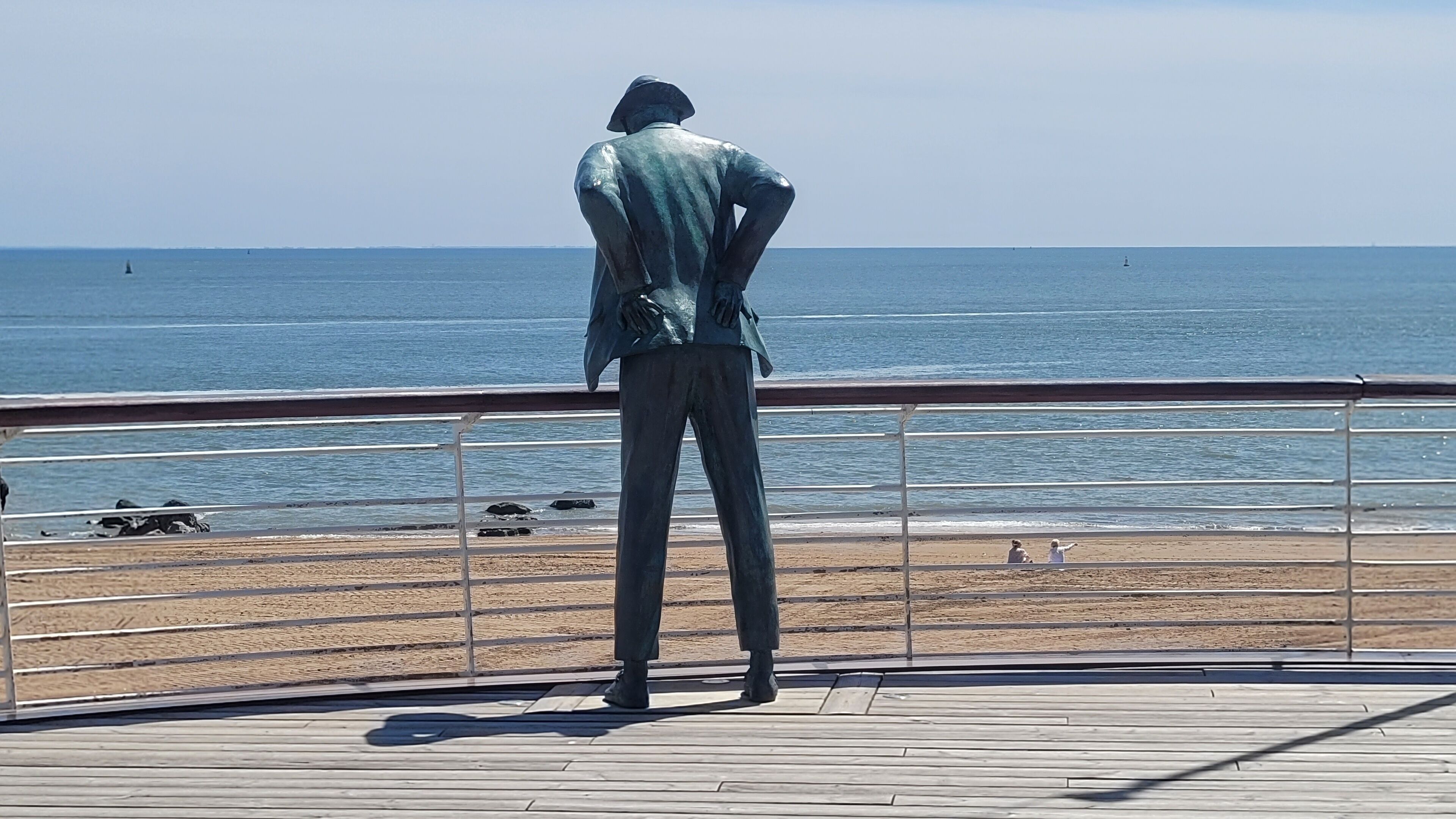 A bronze statue of a man in a suit standing on a wooden boardwalk overlooking the ocean.