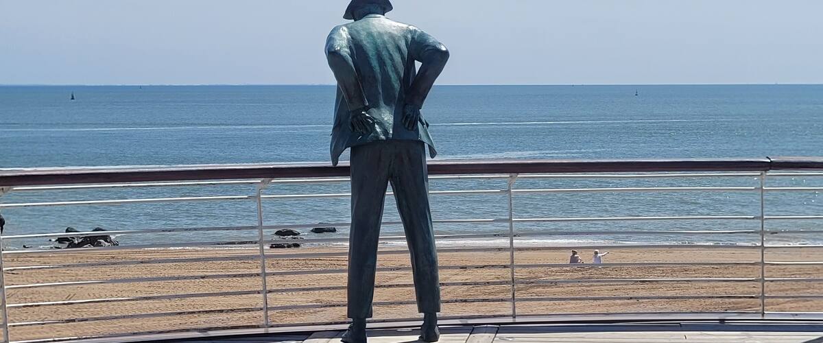 A bronze statue of a man in a suit standing on a wooden boardwalk overlooking the ocean.