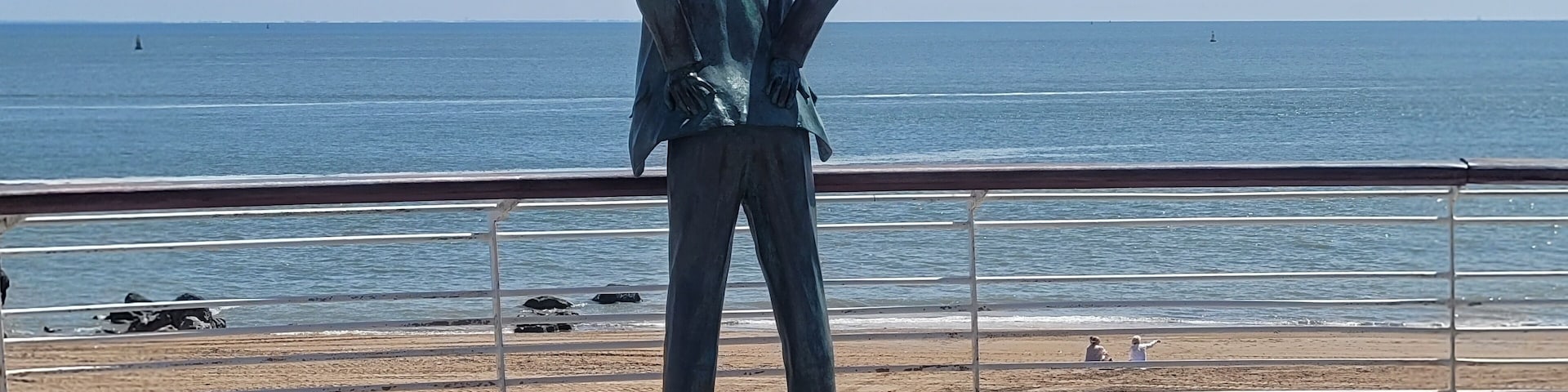 A bronze statue of a man in a suit standing on a wooden boardwalk overlooking the ocean.