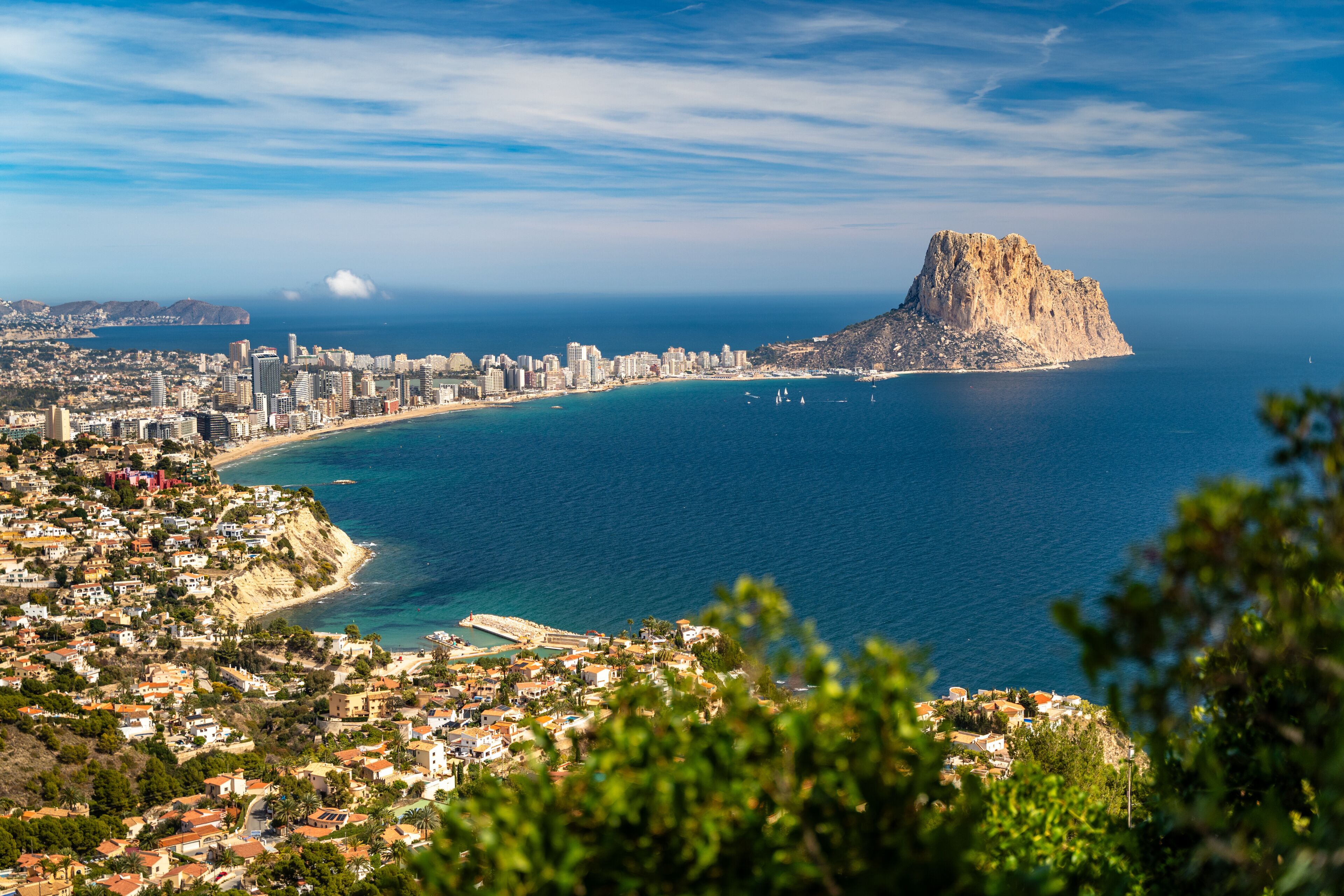 A coastal cityscape with a prominent rocky peninsula, a clear blue sea, and a densely built urban area.
