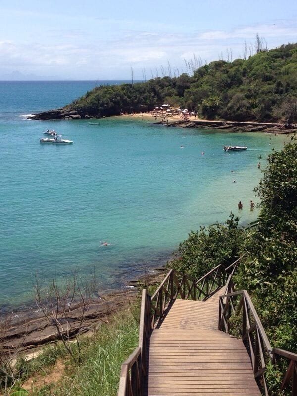 Una escalera de madera que baja a una playa con aguas turquesas cristalinas, embarcaciones y una ladera verde exuberante.