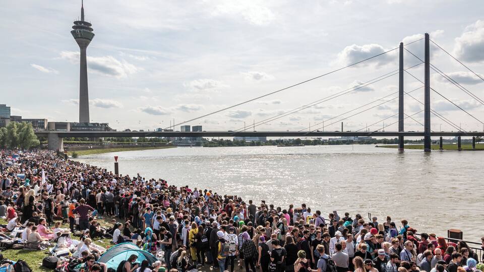 Een drukke rivieroever met een grote menigte, een hoge toren en een brug.