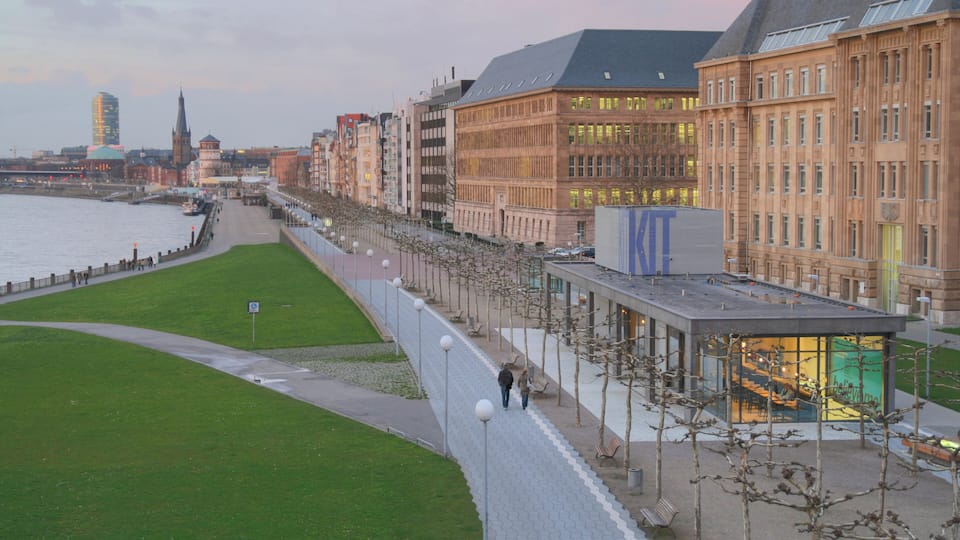 Una passeggiata lungo il fiume con un edificio moderno, una chiesa e un grattacielo sullo sfondo.