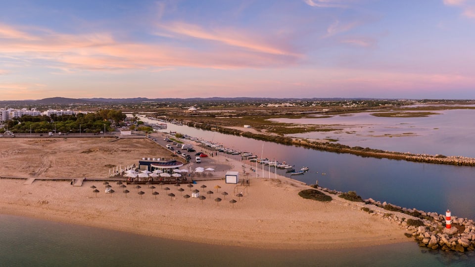 An aerial view of a coastal area with a beach, a pier, boats, and a lighthouse.