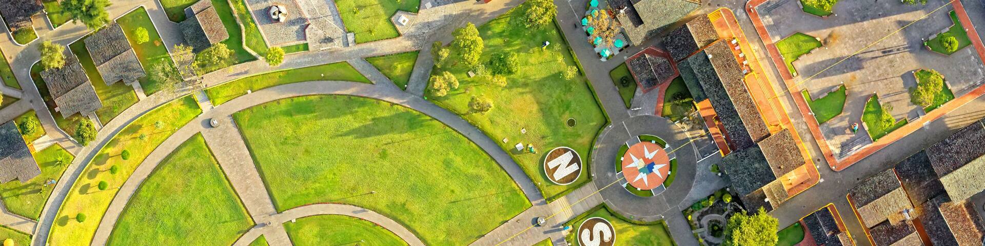 An aerial drone view of the Mitad del Mundo monument at the center of a symmetrical circular park with concentric green lawns in Quito, Ecuador, the equator-line landmark in bright sunlight.