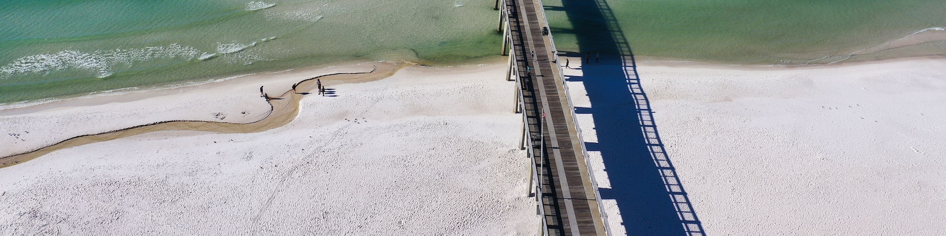 A long pier extending into a clear blue sea with a sandy beach at the end.