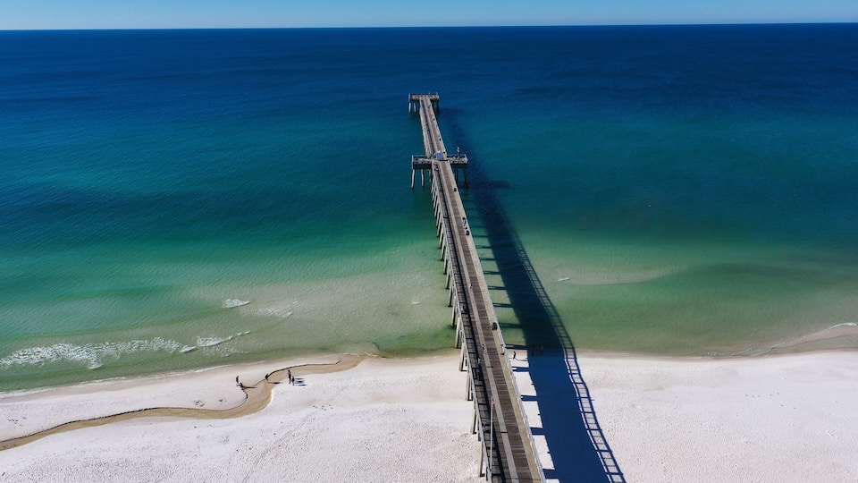 A long pier extending into a clear blue sea with a sandy beach at the end.