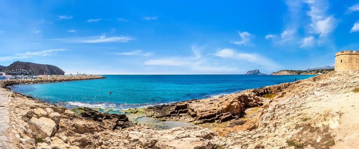 A coastal landscape with a rocky shoreline, a clear blue sea, and a historic tower.