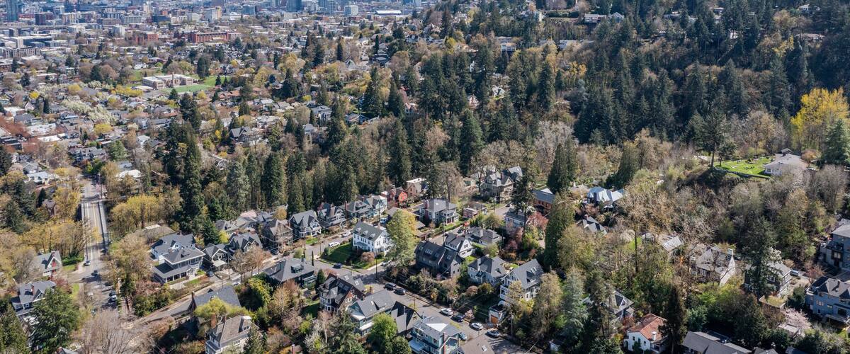Aerial drone view of Northwest Portland, Oregon, showing Forest Park, Nob Hill, and Willamette Heights with greenery, homes, and industrial areas along the Willamette River