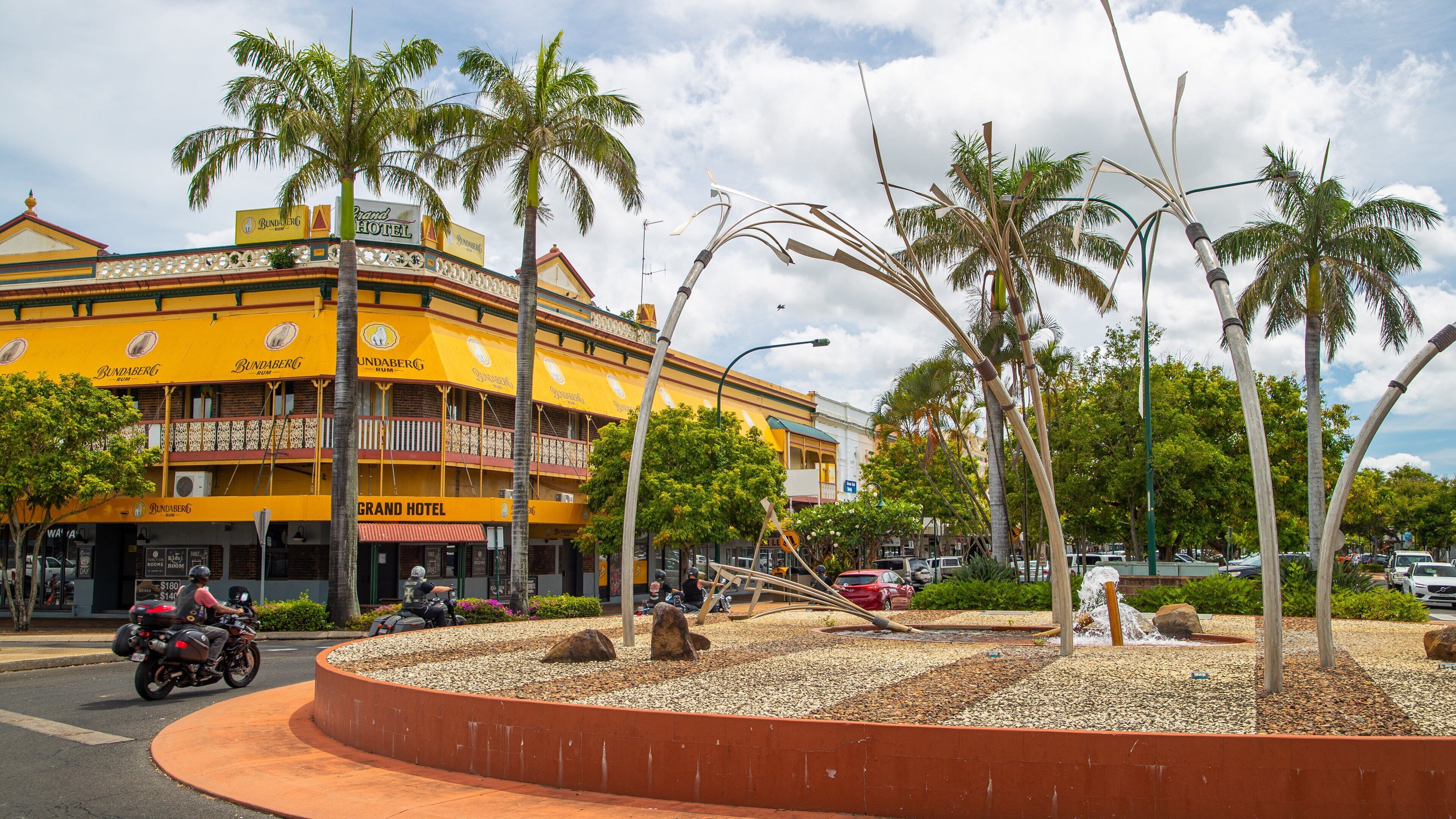 A yellow building with 'Grand Hotel' signage, palm trees, and a roundabout with decorative metal sculptures.