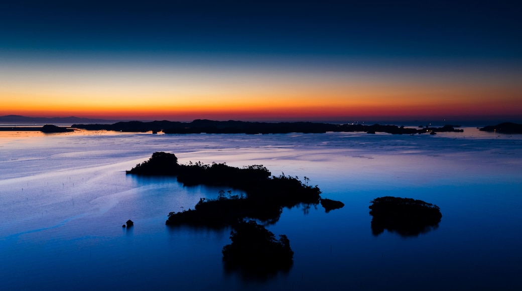 A calm body of water with several dark, rocky formations in the foreground and a sunset in the background.