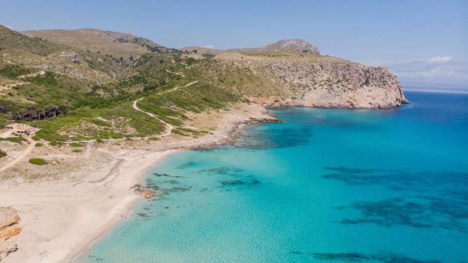 A coastal landscape with a clear turquoise sea, a sandy beach, and a winding path leading up a green hillside.